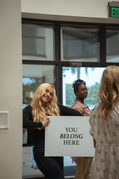 A smiling woman holding a You Belong Here sign near the conference entrance.