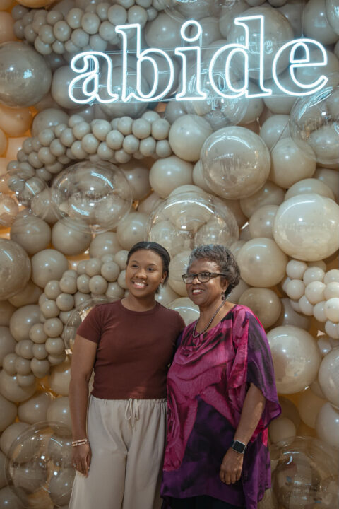 Two women smiling in front of a glowing abide neon sign and a wall of soft balloons.